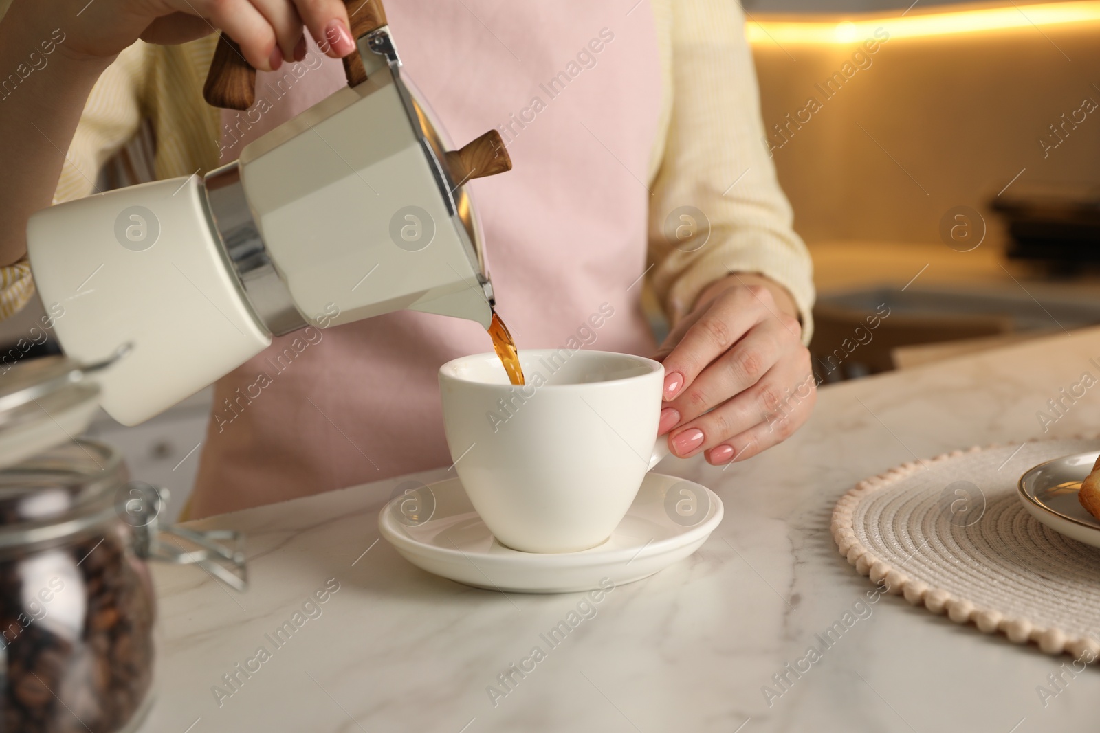 Photo of Woman pouring aromatic coffee from moka pot into cup at white marble table, closeup