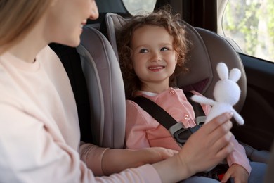 Photo of Cute little girl sitting in child safety seat near mother with toy rabbit inside car