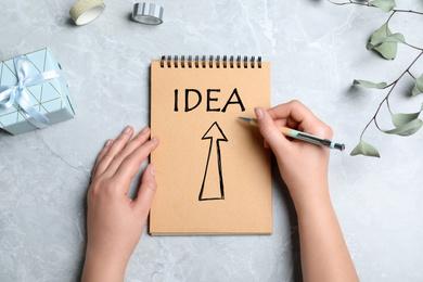 Woman writing word IDEA in notebook at grey table, top view