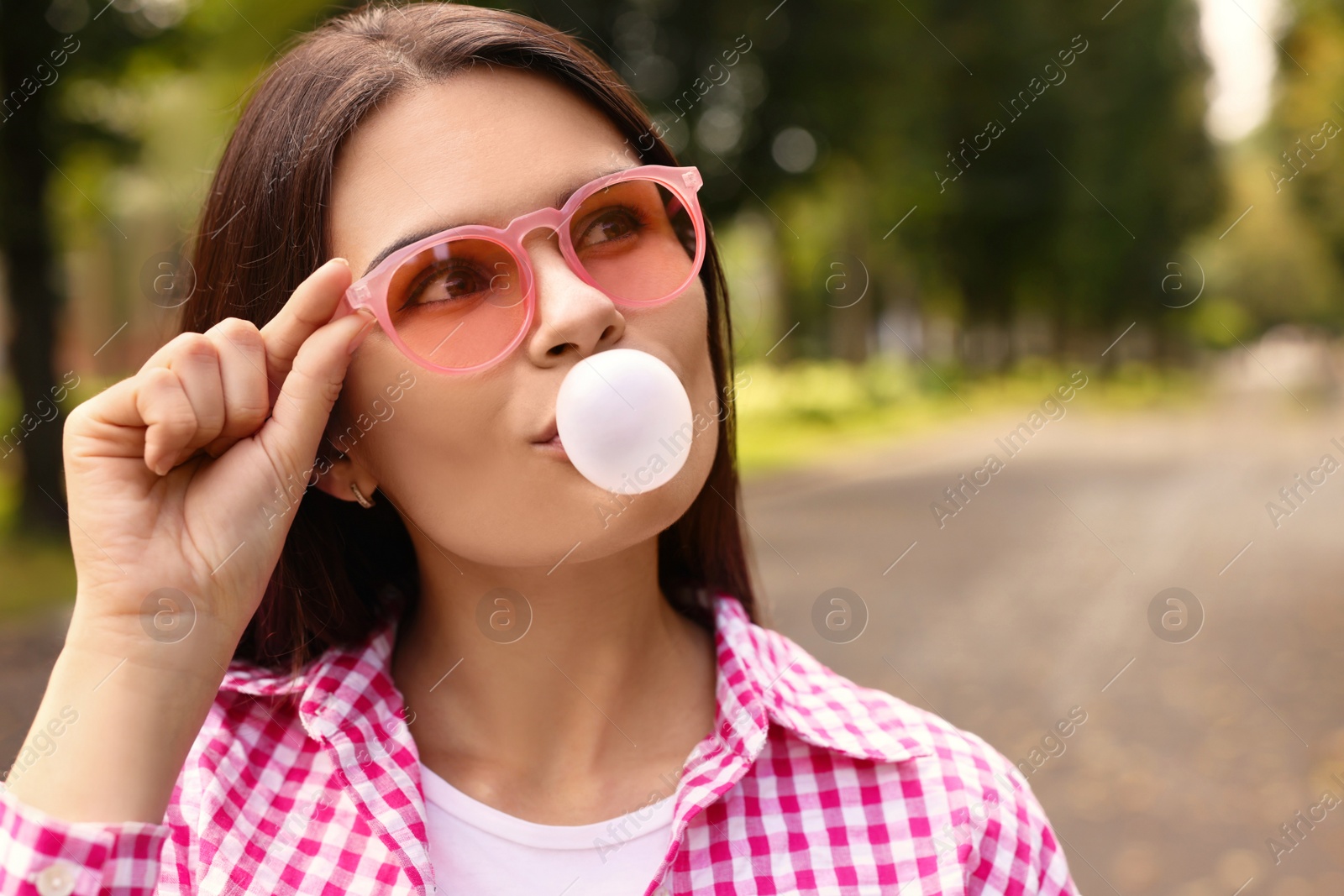 Photo of Beautiful young woman with pink sunglasses blowing chewing gum outdoors, space for text