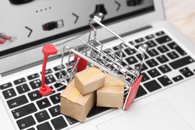 Photo of Internet store. Small cardboard boxes and shopping cart on laptop, closeup