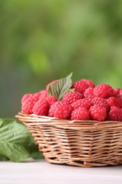 Wicker basket with tasty ripe raspberries and leaves on white wooden table against blurred green background, closeup. Space for text