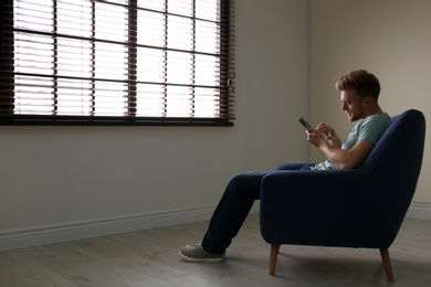 Lonely man with mobile phone in armchair near window indoors