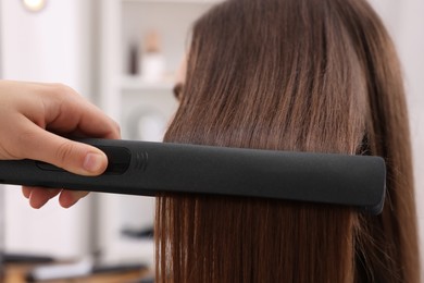 Photo of Hairdresser straightening woman's hair with flat iron in salon, closeup