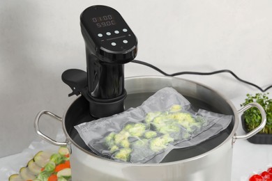 Photo of Sous vide cooker and vacuum packed broccoli in pot on white table. Thermal immersion circulator