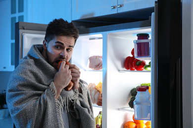 Young man eating sausages near open refrigerator at night