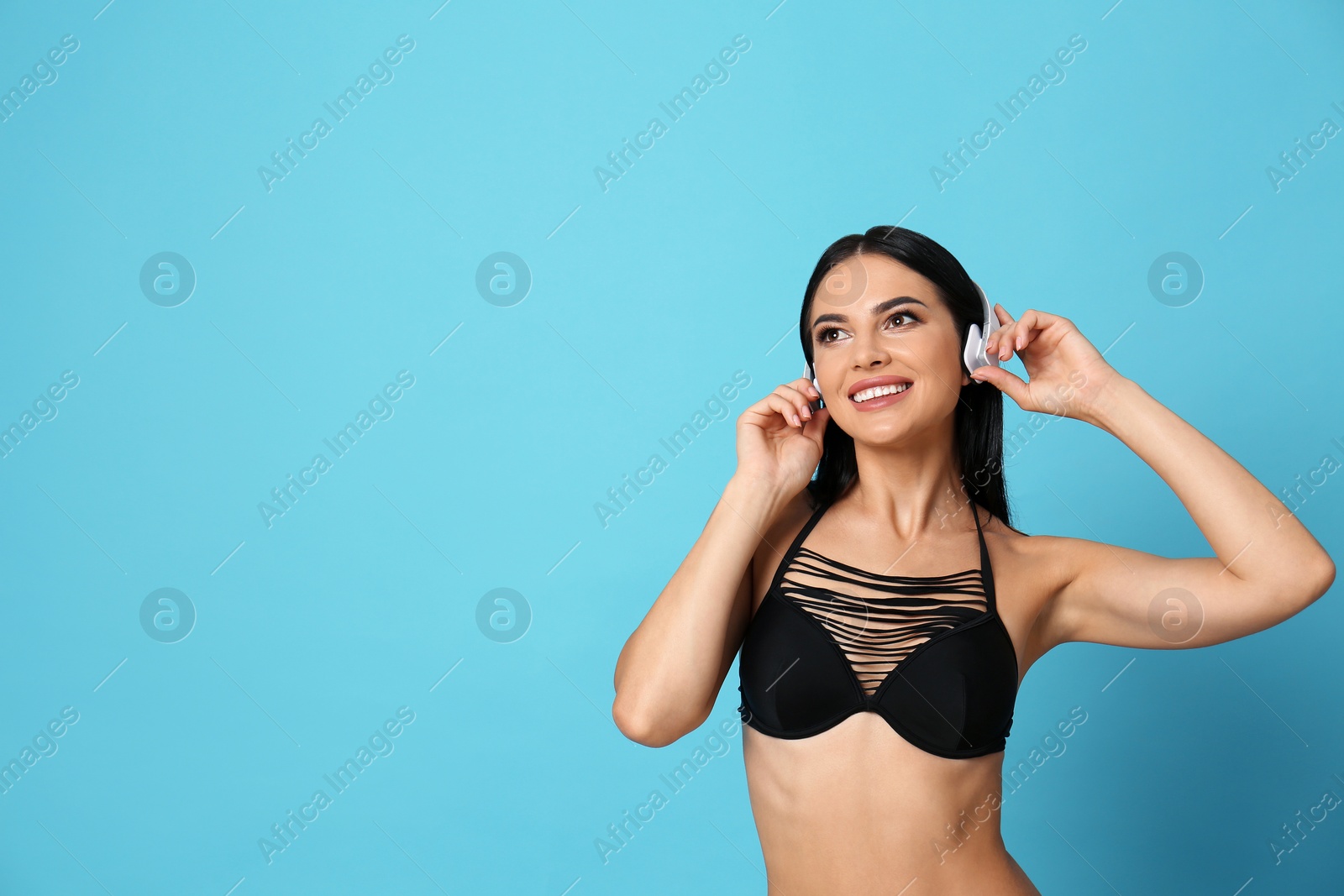 Photo of Beautiful young woman in black bikini with headphones on light blue background. Space for text