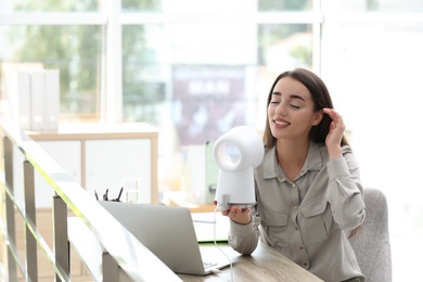 Photo of Young woman enjoying air flow from portable fan at workplace, space for text. Summer heat