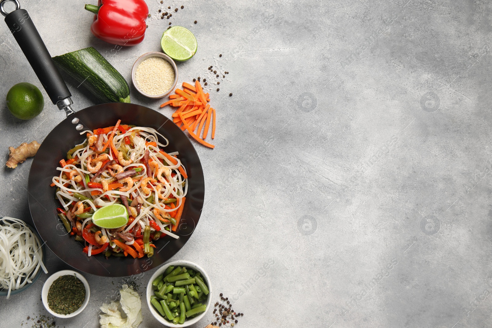 Photo of Shrimp stir fry with noodles and vegetables in wok surrounded by ingredients on grey table, flat lay. Space for text