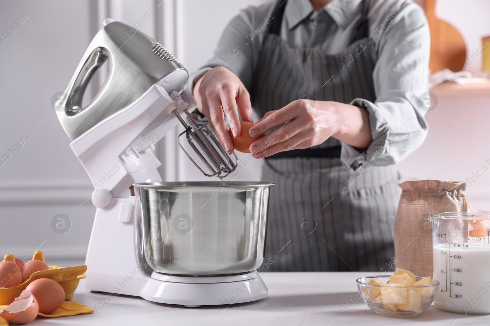 Photo of Woman adding egg into bowl of stand mixer while making dough at table indoors, closeup