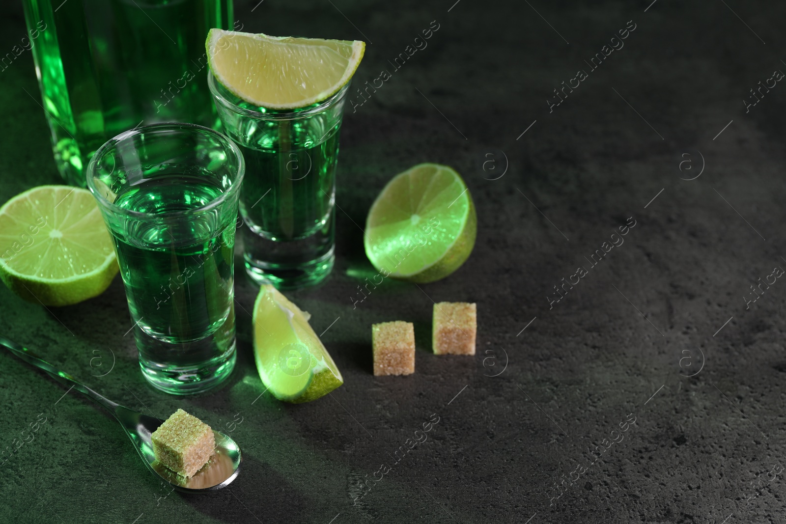 Photo of Absinthe in shot glasses, spoon, lime and brown sugar cubes on gray textured table, space for text. Alcoholic drink