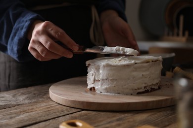 Photo of Woman smearing sponge cake with cream at wooden table, closeup