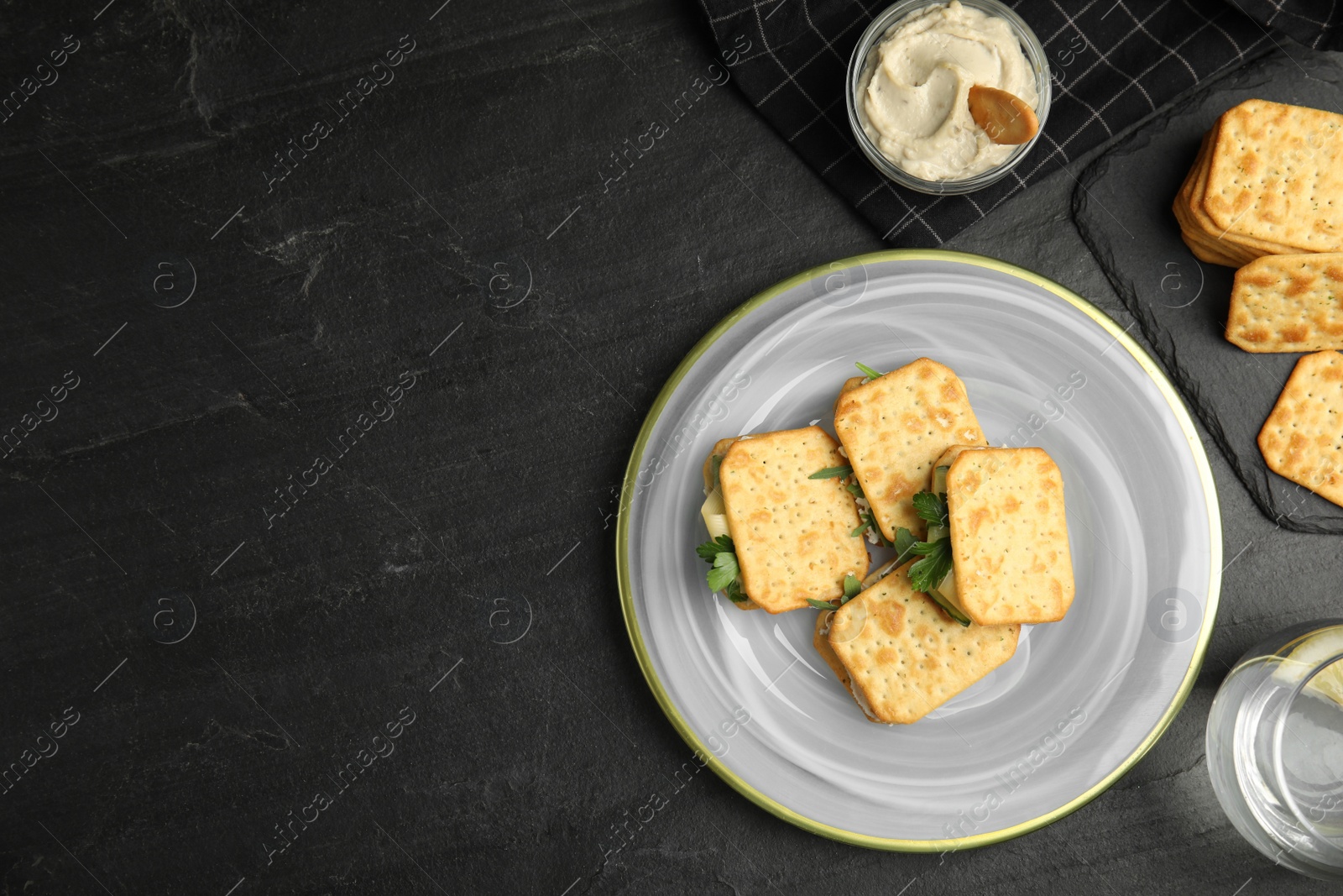 Photo of Delicious crackers with cream cheese, cucumber and parsley on black table, flat lay. Space for text