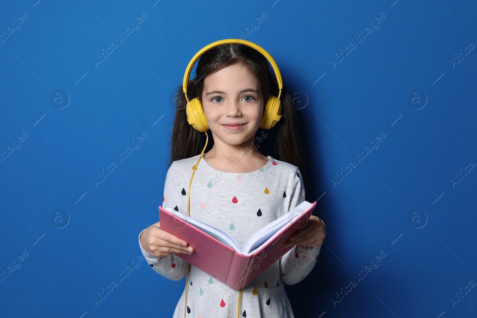 Photo of Cute little girl with headphones listening to audiobook on blue background