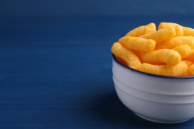 Photo of Bowl of tasty cheesy corn puffs on blue wooden table, space for text
