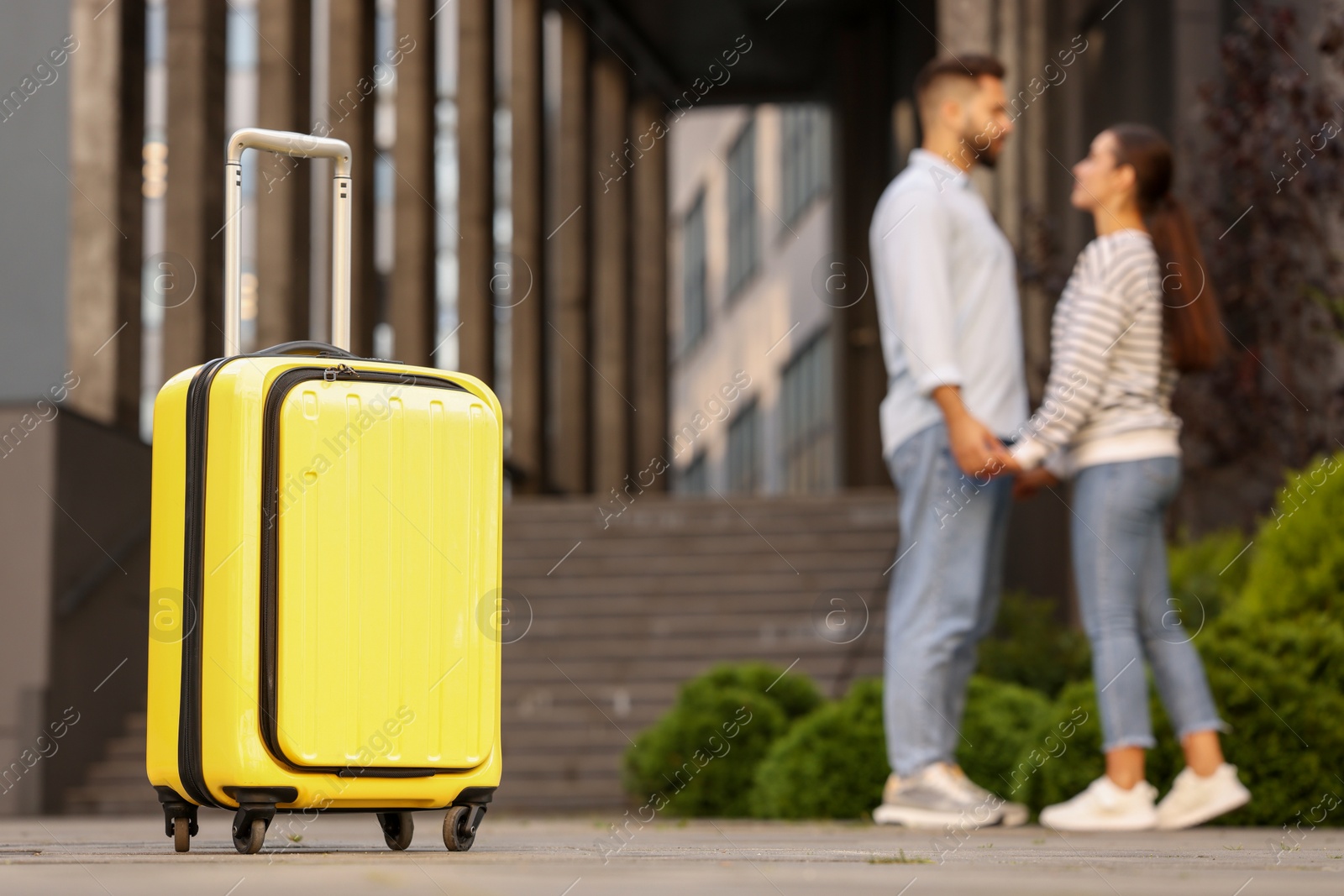 Photo of Long-distance relationship. Young couple holding hands near building outdoors, focus on yellow suitcase