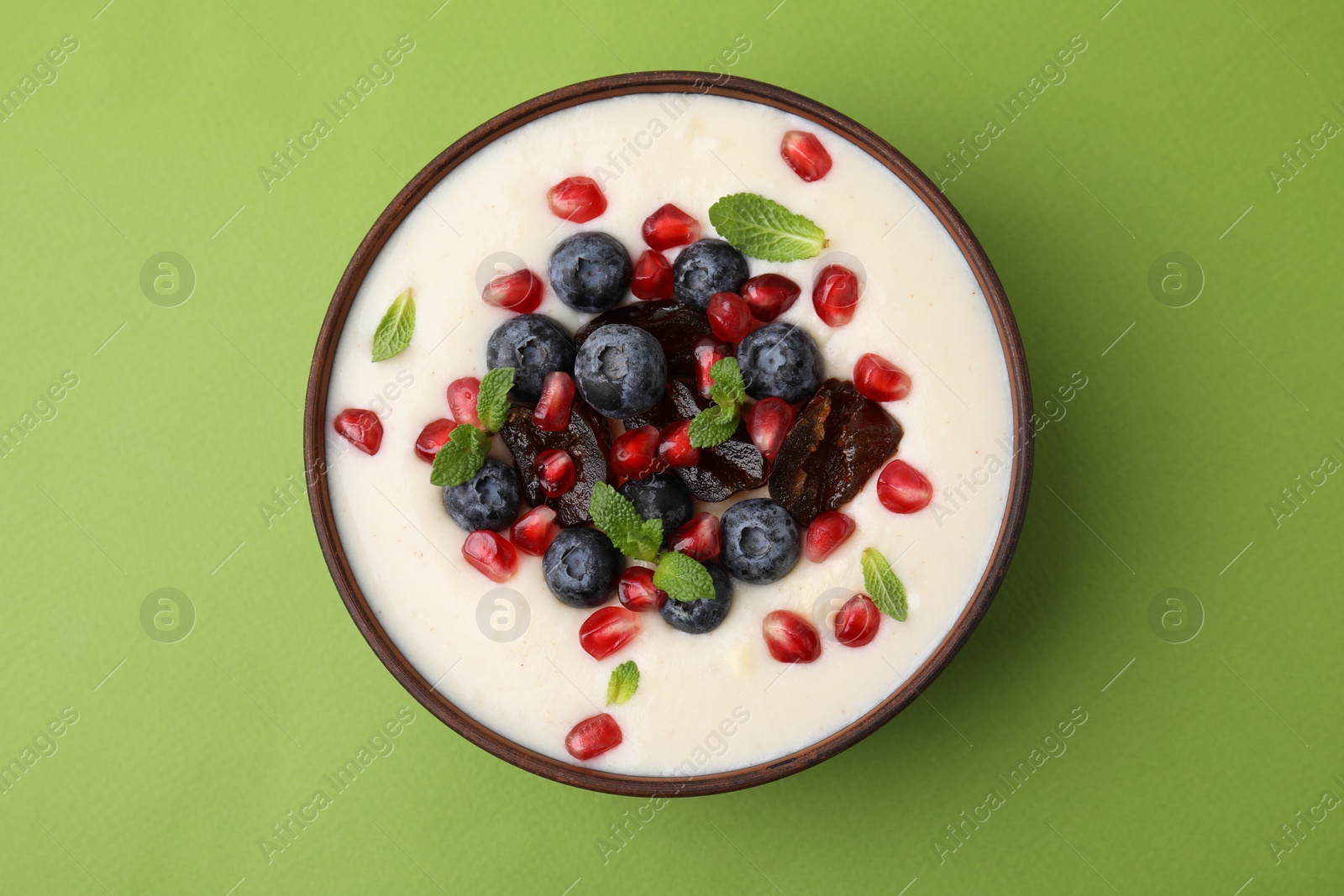 Photo of Delicious semolina pudding with blueberries, pomegranate, dates and mint in bowl on green table, top view