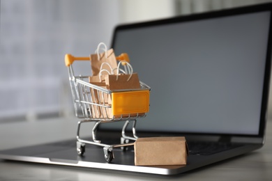 Photo of Internet shopping. Modern laptop and small cart with bags and box on table indoors