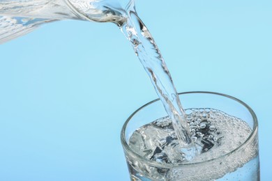 Pouring water from jug into glass on light blue background, closeup