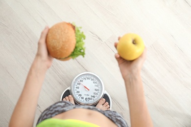 Woman holding tasty sandwich and fresh apple while measuring her weight on floor scales, top view. Choice between diet and unhealthy food