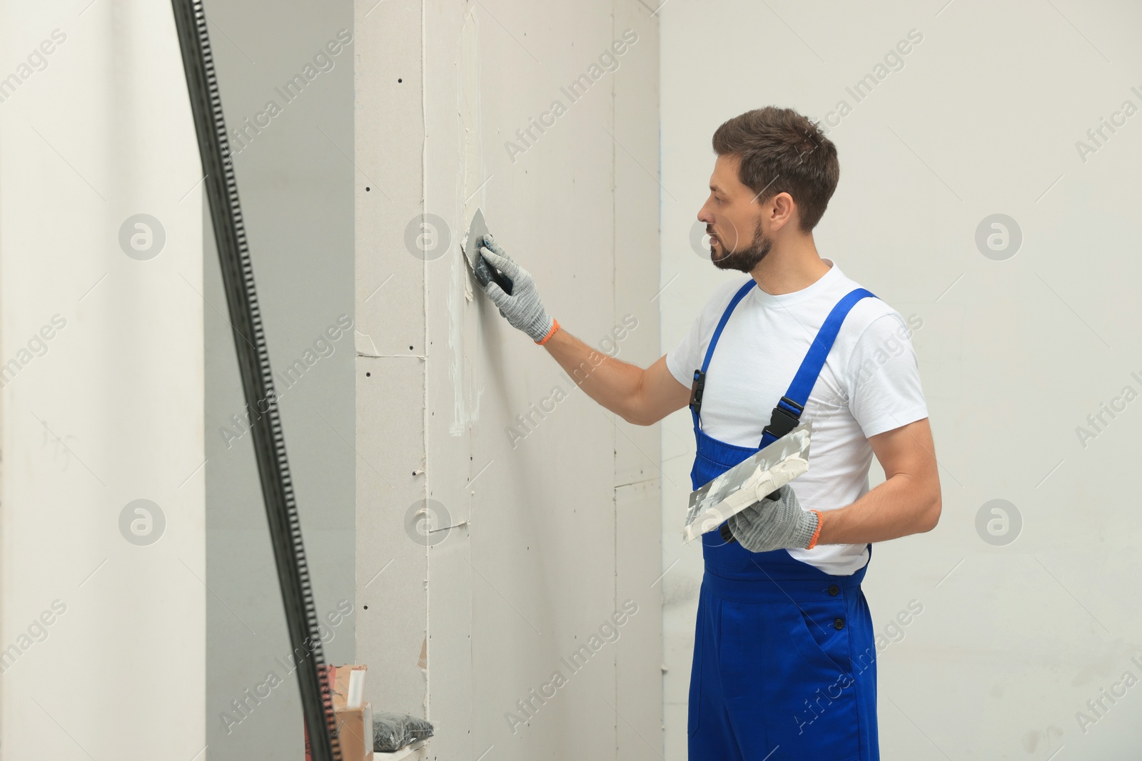 Photo of Professional worker in uniform plastering wall with putty knife indoors