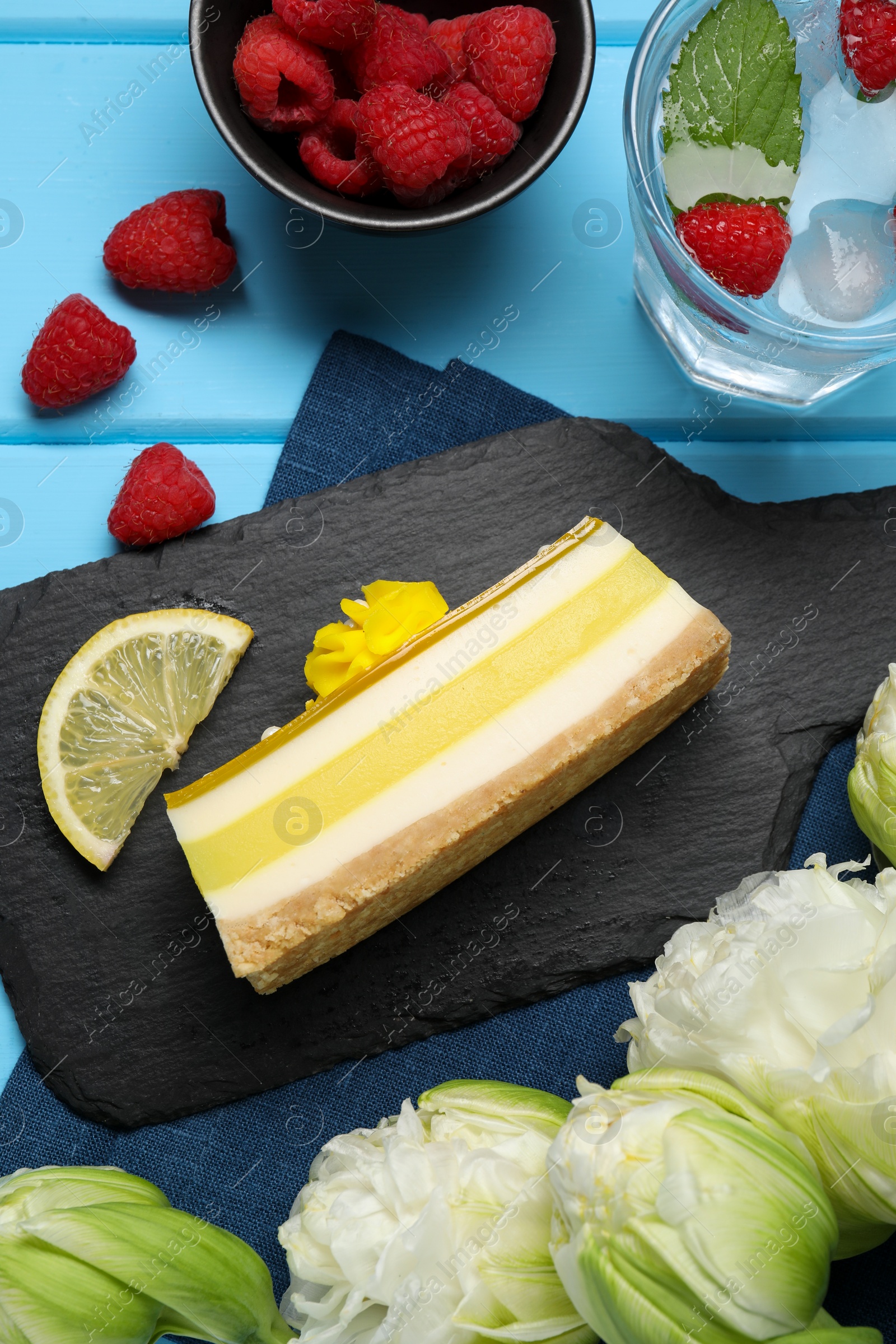 Photo of Delicious cheesecake with lemon, raspberries and peonies on light blue wooden table, flat lay