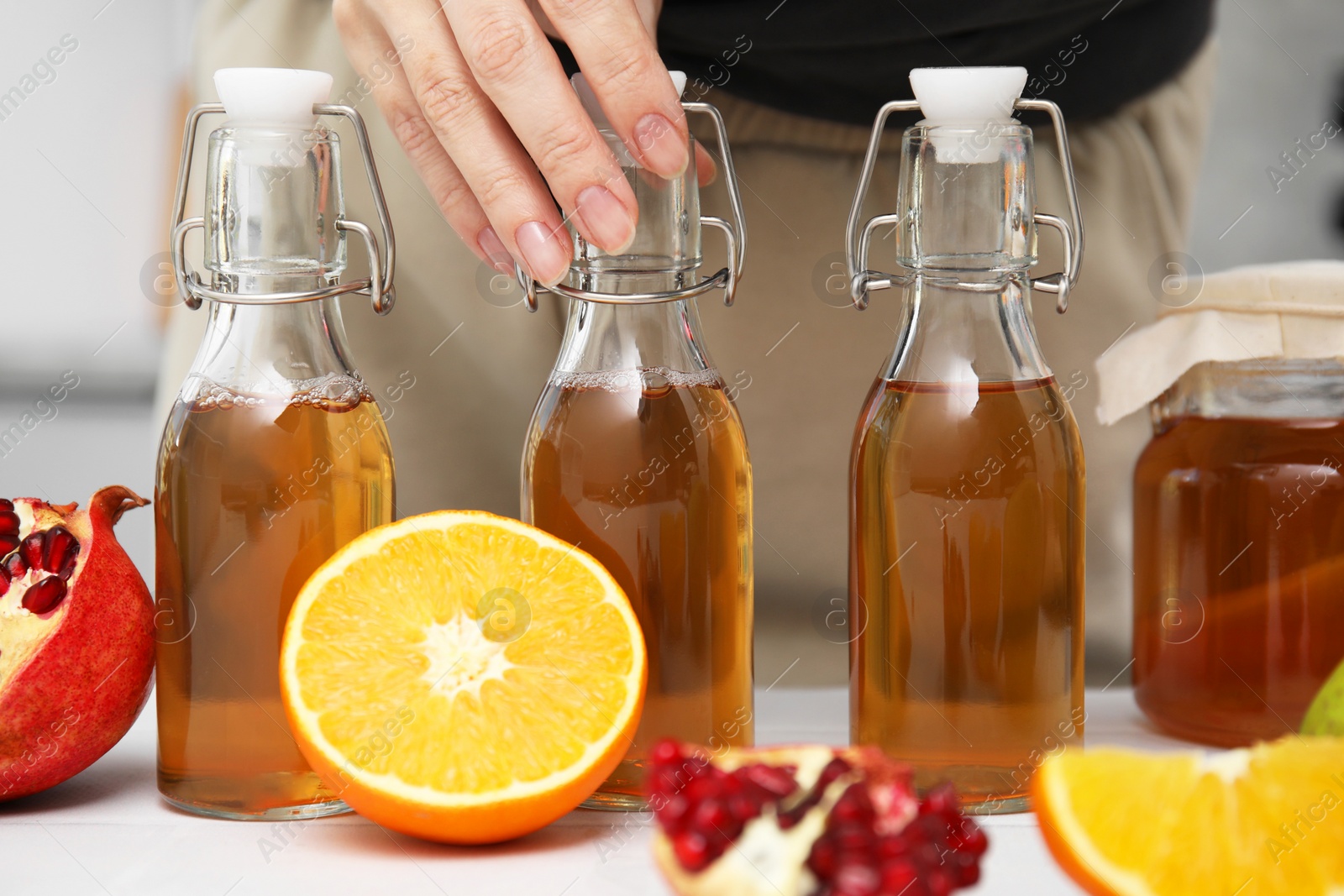 Photo of Woman taking bottle of fermented kombucha at white table indoors, closeup