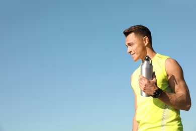 Young sporty man with water bottle against blue sky on sunny day. Space for text