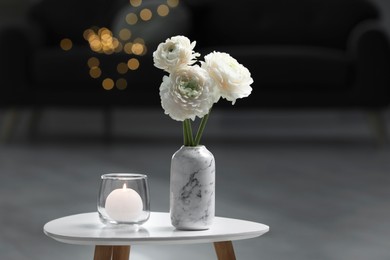 Vase with beautiful white flowers and burning candle on table in room, bokeh effect. Stylish interior design