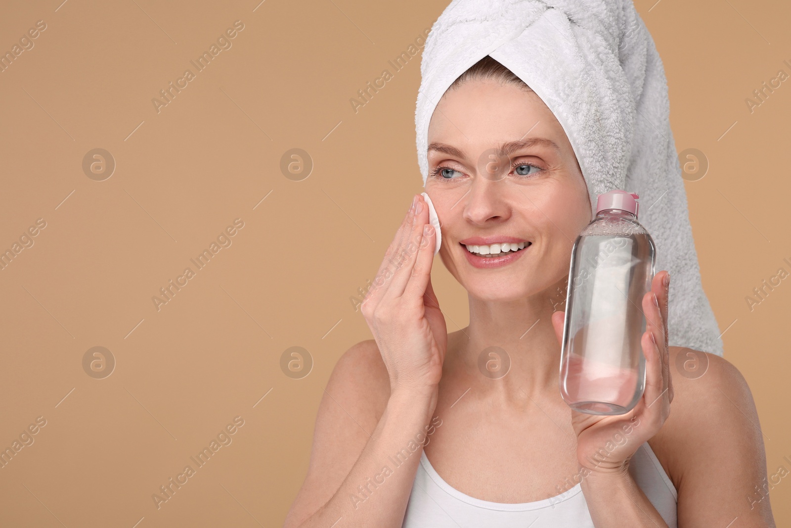 Photo of Beautiful woman in terry towel removing makeup with cotton pad on beige background, space for text