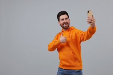 Photo of Smiling man taking selfie with smartphone and showing thumbs up on grey background, space for text