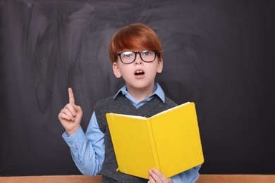 Cute schoolboy in glasses with book near blackboard