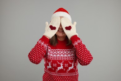 Happy senior woman in Christmas sweater and Santa hat covering her face with hands in knitted mittens on grey background