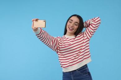 Smiling young woman taking selfie with smartphone on light blue background, space for text