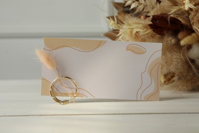 Blank invitation card, ring, dry leaves and flowers on white wooden table