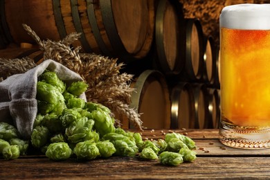 Glass of tasty light beer, fresh hops and wheat spikes on wooden table in cellar