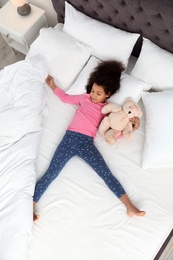 Photo of Cute little African-American girl with teddy bear sleeping in bed, top view