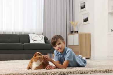 Photo of Little boy brushing cute ginger cat's fur on soft carpet at home