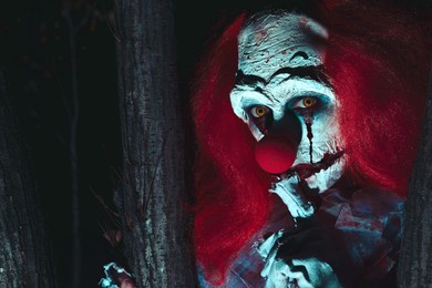 Photo of Terrifying clown hiding behind trees outdoors at night. Halloween party costume