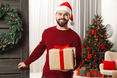 Photo of Young man in Santa hat with Christmas gift box indoors. Sending present by mail