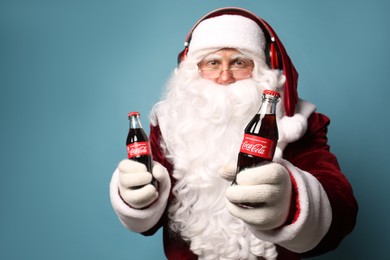 MYKOLAIV, UKRAINE - JANUARY 18, 2021: Santa Claus holding Coca-Cola bottles and listening to music with headphones on light blue background