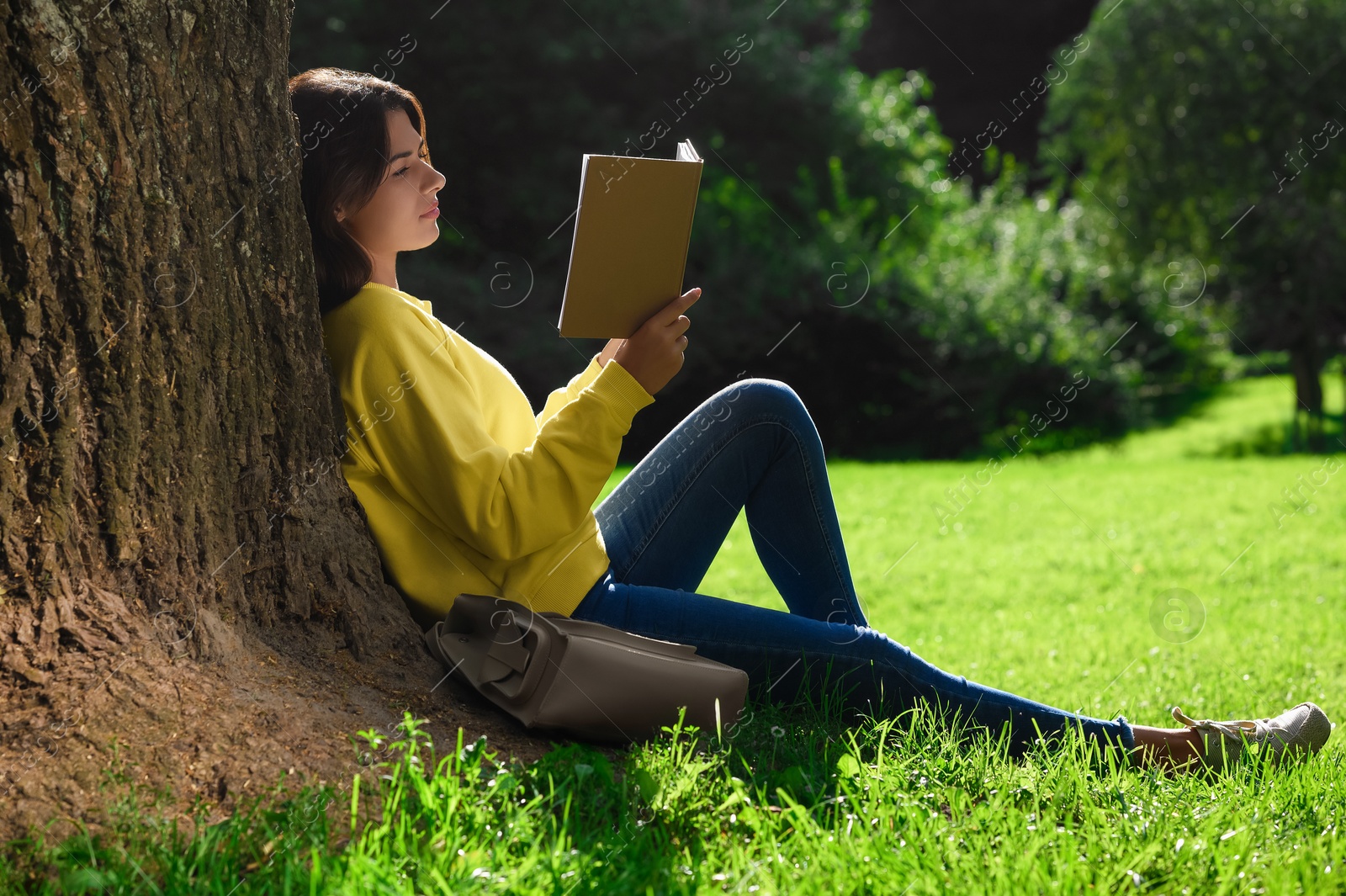 Photo of Young woman reading book near tree in park on sunny day