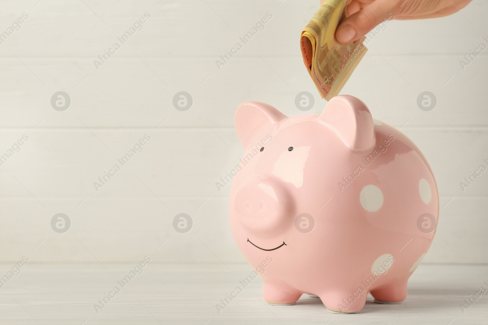 Photo of Woman putting banknotes into piggy bank at white wooden table, closeup. Space for text