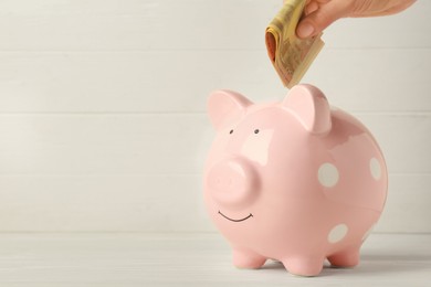 Woman putting banknotes into piggy bank at white wooden table, closeup. Space for text