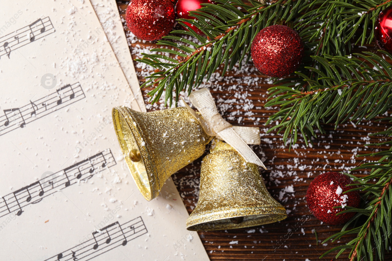 Photo of Golden shiny bells with bow, music sheet and red decorative balls on wooden table, flat lay. Christmas decoration