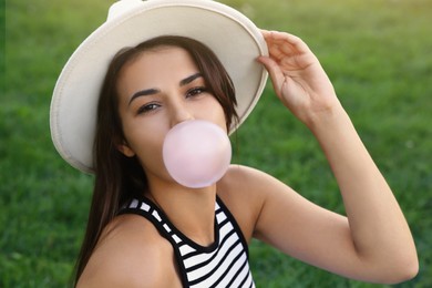 Photo of Beautiful woman in hat blowing gum on green grass outdoors