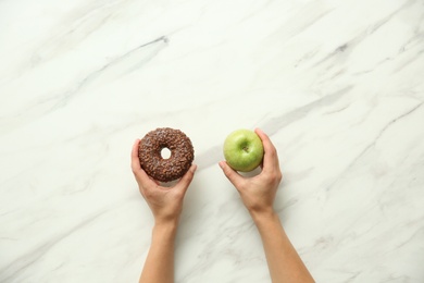 Top view of woman choosing between doughnut and healthy apple on white marble table, closeup