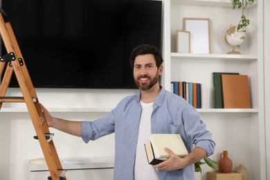 Happy man with books near wooden folding ladder indoors