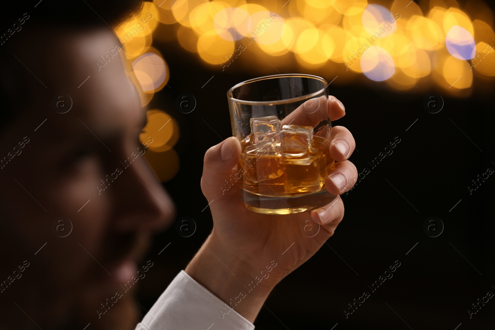 Photo of Man holding glass of whiskey with ice cubes against blurred lights, selective focus