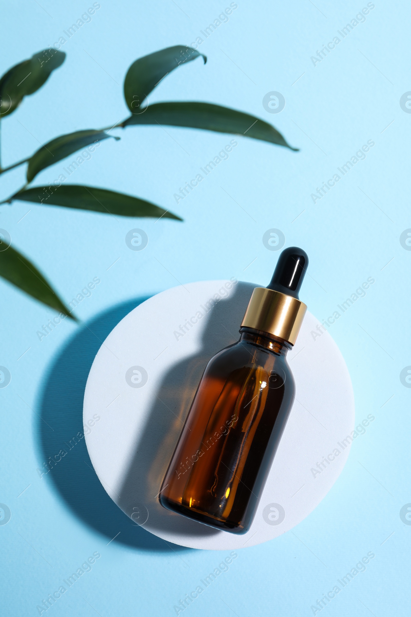 Photo of Bottle with cosmetic oil on podium and green leaves on light blue background, flat lay